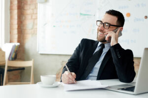 A male Sales Manager sitting at a computer and talking on the phone.