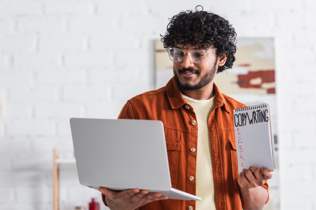 Mężczyzna w prawej ręce trzyma laptop i spogląda na niego, a w lewej notes z napisem "Copywriting" i listą punktów do uzupełnienia.